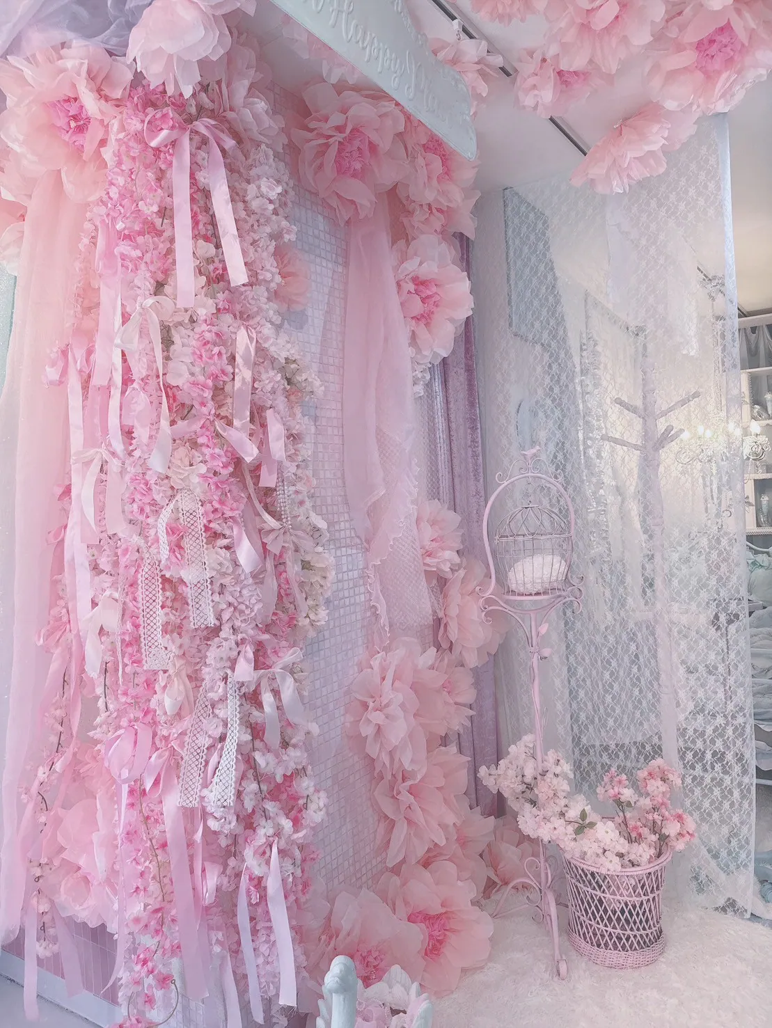 ピンクの造花とリボンで埋め尽くされた壁が印象的なかわいい撮影ブース。白いレースカーテンや鳥かごスタンド、桜の花を挿したバスケットを配置